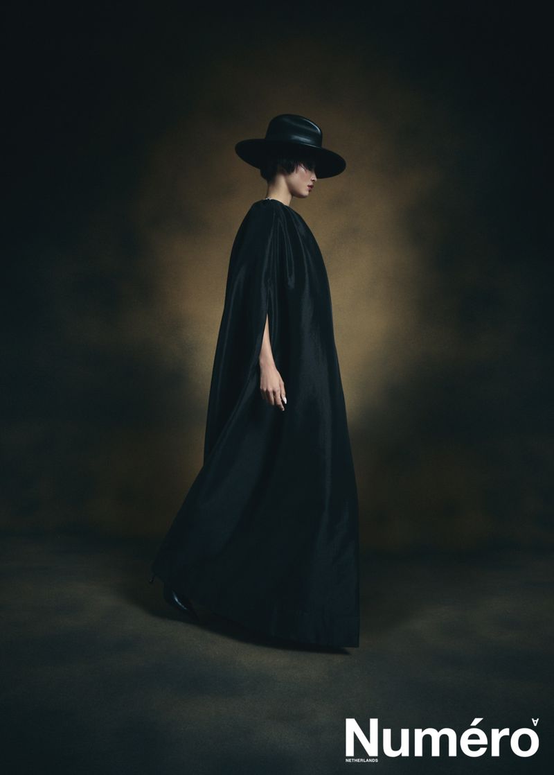 Editorial fashion shot: Elegant woman in a full-length black dress and wide-brimmed hat poses in a studio with a mottled background.