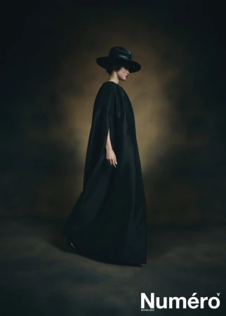 Editorial fashion shot: Elegant woman in a full-length black dress and wide-brimmed hat poses in a studio with a mottled background.