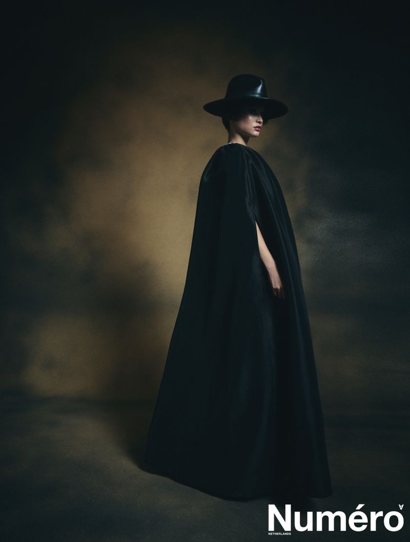 A woman in a black dress and hat strikes a pose in a studio with a dark background for an elegant fashion editorial shot.