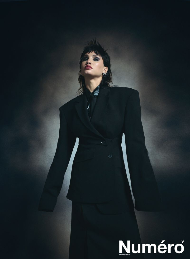 Fashion editorial shot of a woman with a mullet wearing a black oversized blazer and skirt, standing in front of a dark backdrop.