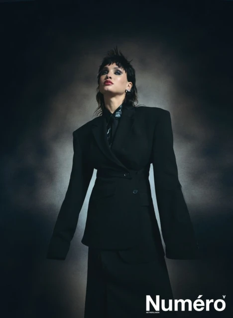 Fashion editorial shot of a woman with a mullet wearing a black oversized blazer and skirt, standing in front of a dark backdrop.