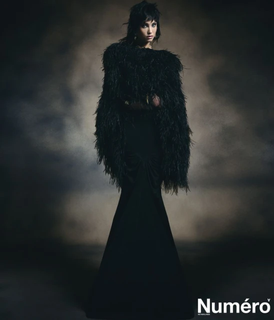 Adult woman in a dramatic full-length black gown with a feather boa in a dark, hazy studio environment for a fashion editorial.
