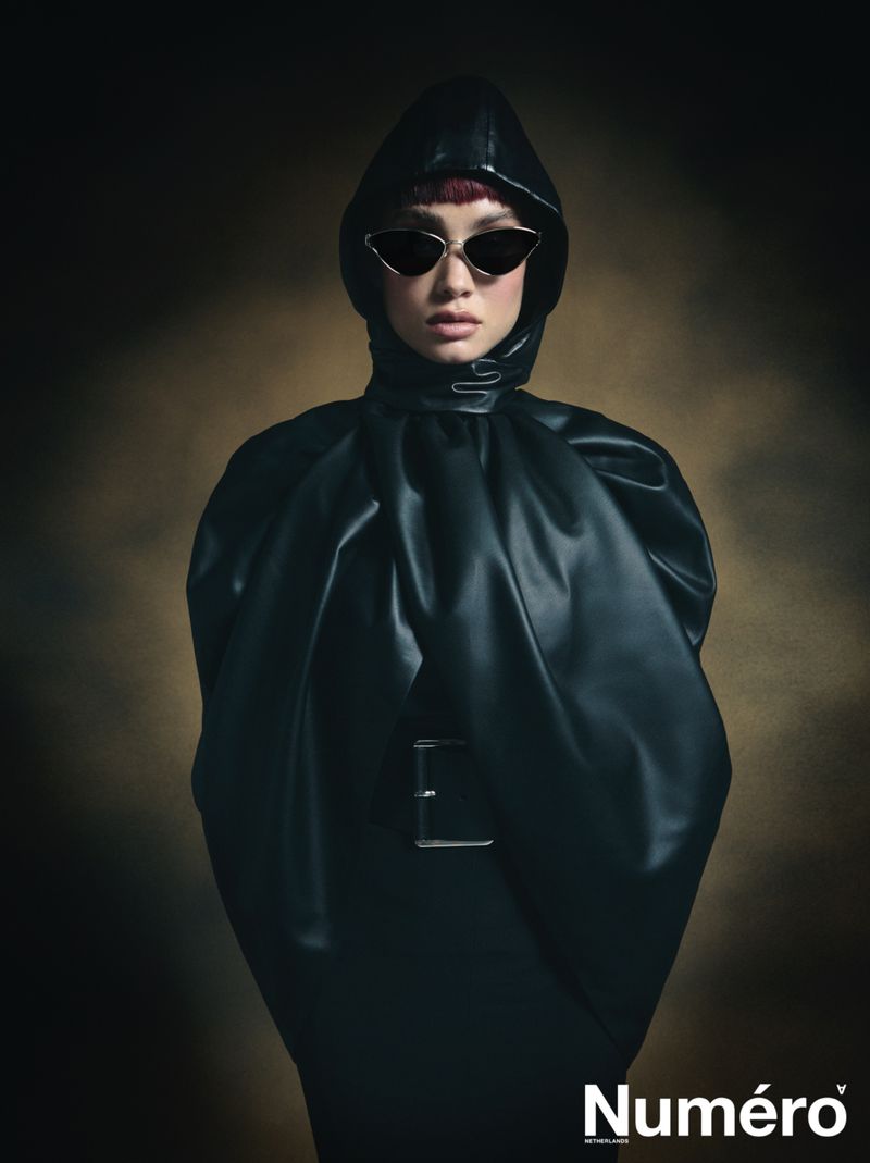 Portrait of a fashionable person wearing sunglasses and a dark, hooded outfit against a diffused brown background with the brand name visible.