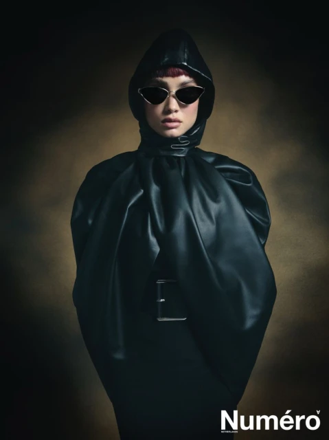 Portrait of a fashionable person wearing sunglasses and a dark, hooded outfit against a diffused brown background with the brand name visible.