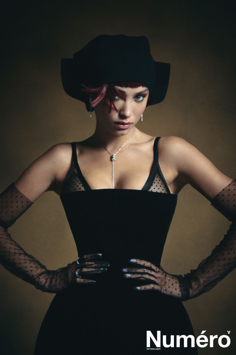 Model Rose Bertram posing with hands on hips wearing a black corset dress, sheer polka dot opera gloves, and a sculptural black hat.