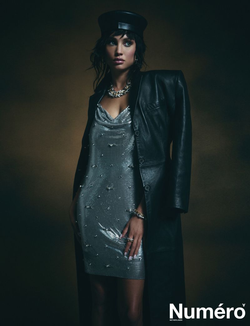 A woman in a shiny silver dress and black leather coat and cap poses in front of a neutral background for a fashion editorial.