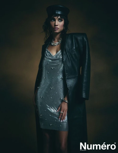 A woman in a shiny silver dress and black leather coat and cap poses in front of a neutral background for a fashion editorial.