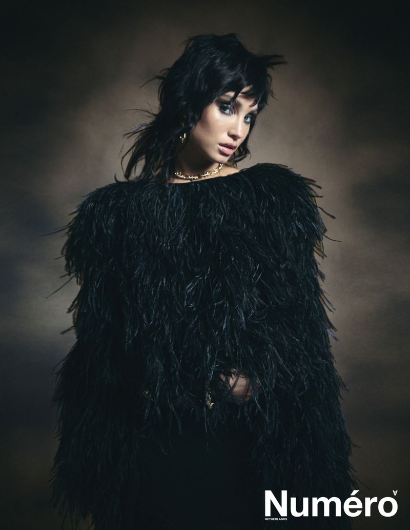 Portrait of a young woman with dark hair wearing a black feathered top against a blurred brown background in a fashion editorial shot.