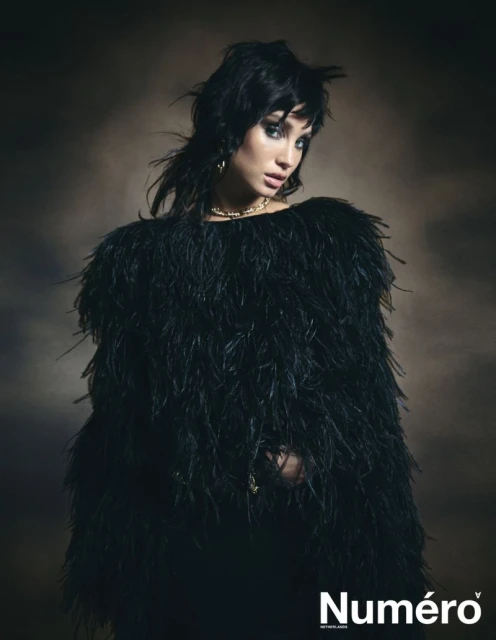 Portrait of a young woman with dark hair wearing a black feathered top against a blurred brown background in a fashion editorial shot.