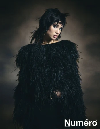 Portrait of a young woman with dark hair wearing a black feathered top against a blurred brown background in a fashion editorial shot.