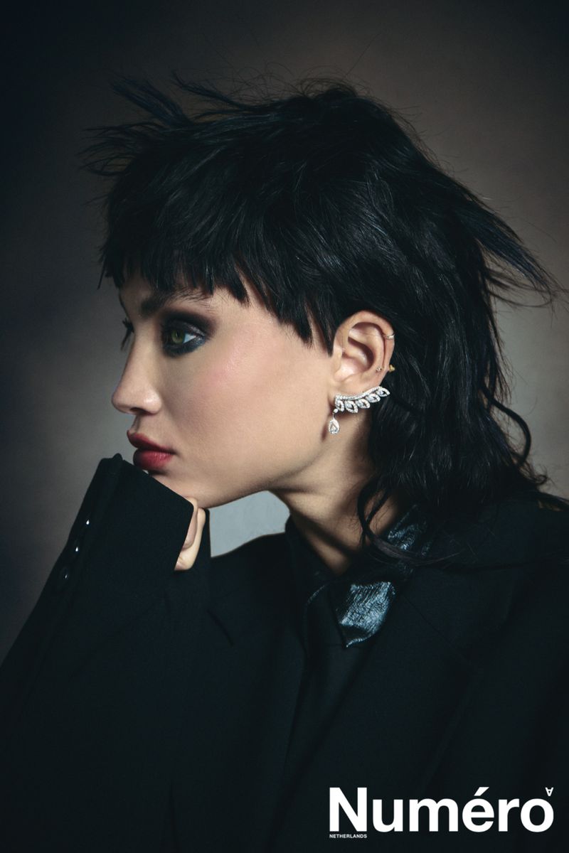 Close-up of a woman with dark hair and smokey eyes wearing a black jacket and statement earring, posing in a studio. Editorial fashion.