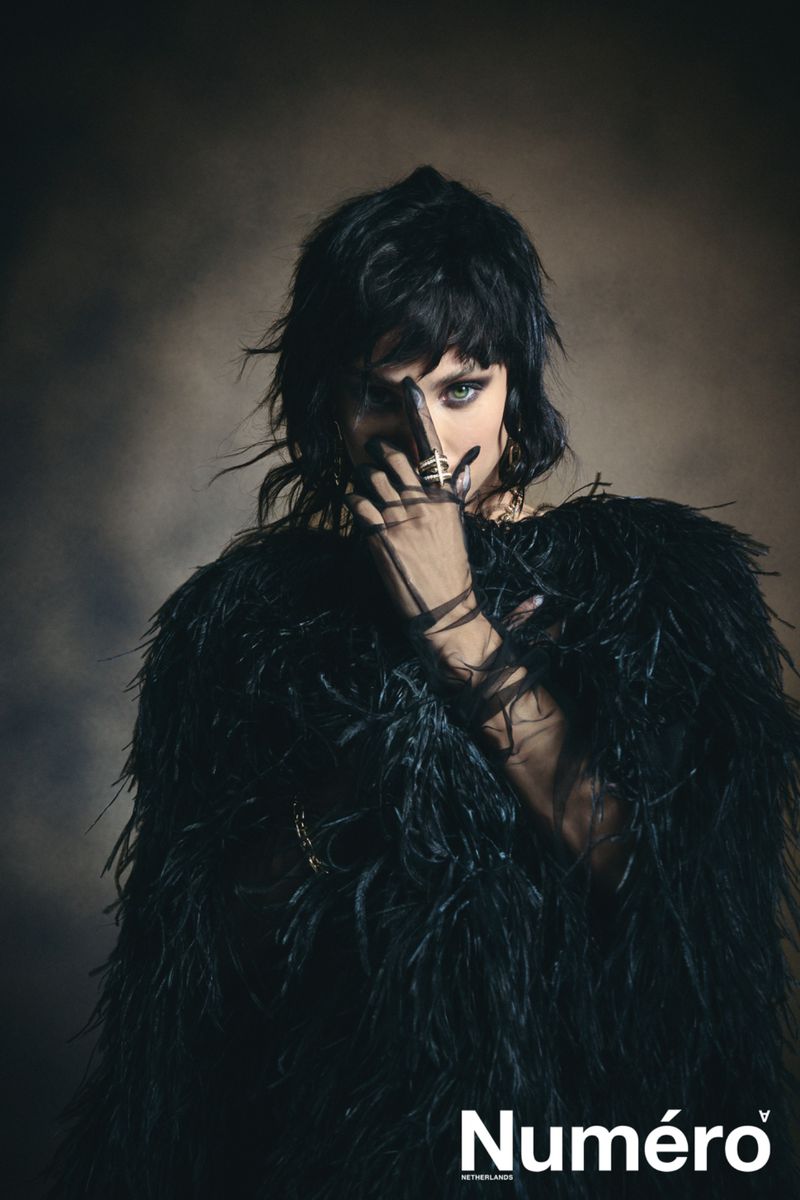 Editorial portrait of a woman with dark hair, wearing a black feathered garment and jewelry, covering her face. 'Numéro' logo visible.