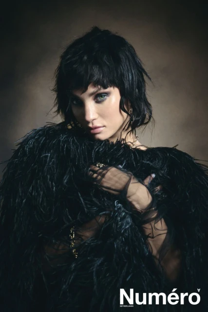 A portrait of an adult female with short black hair and dramatic eye makeup, wearing a feathered coat against a diffused, neutral background.