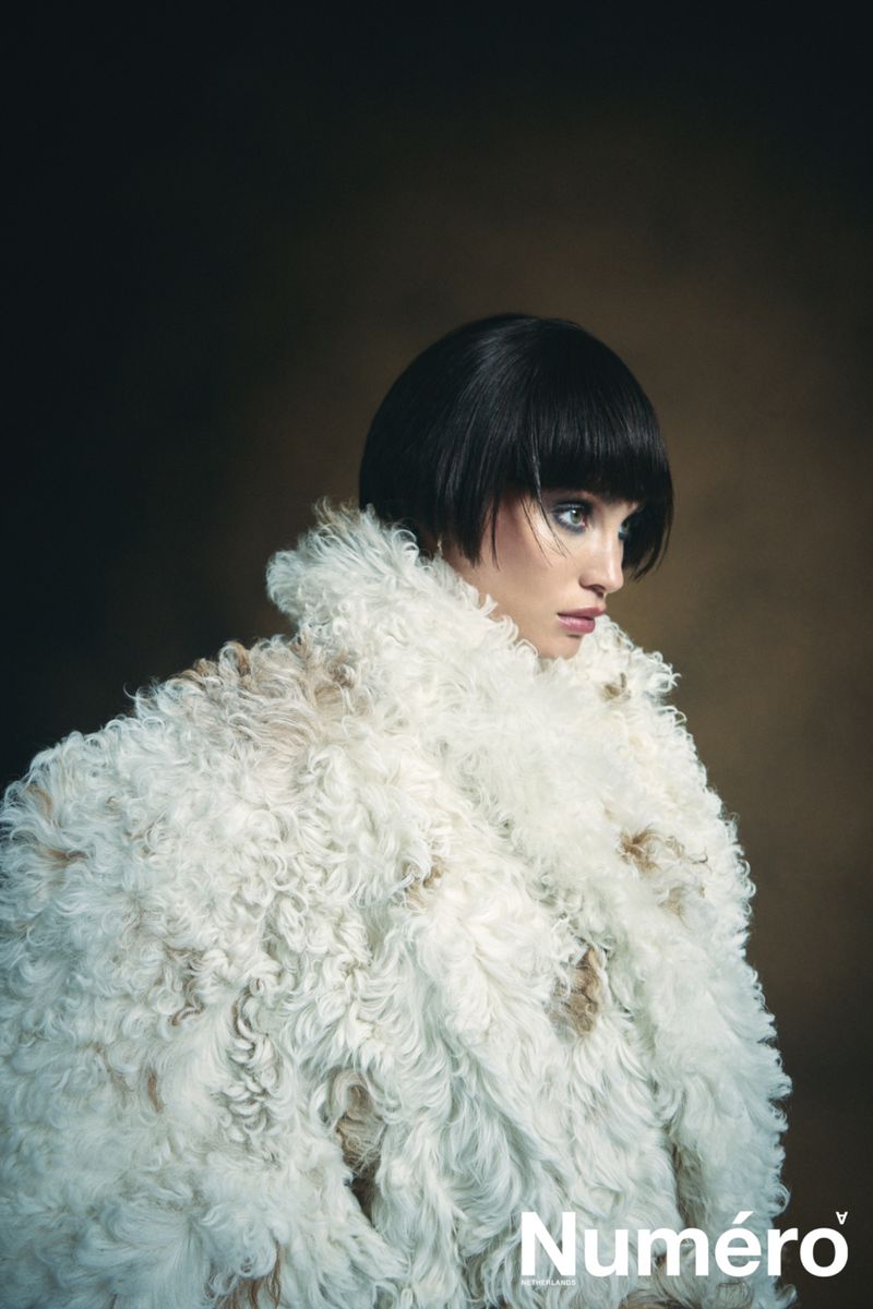 Fashion portrait of a woman with a blunt bob hairstyle wearing a large white faux fur coat, set against a dark brown studio backdrop.