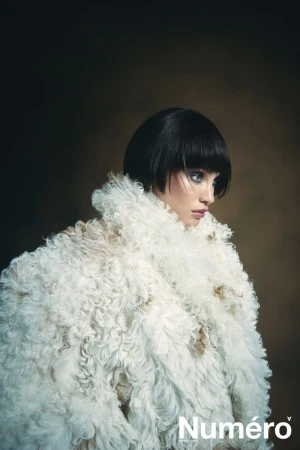 Fashion portrait of a woman with a blunt bob hairstyle wearing a large white faux fur coat, set against a dark brown studio backdrop.