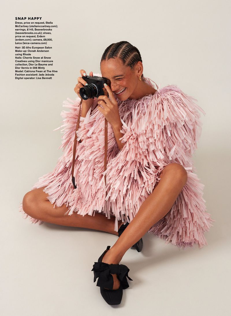 Model Catriona Frean seated cross-legged in a pink fringe dress and black bow heels while holding a camera to her face.