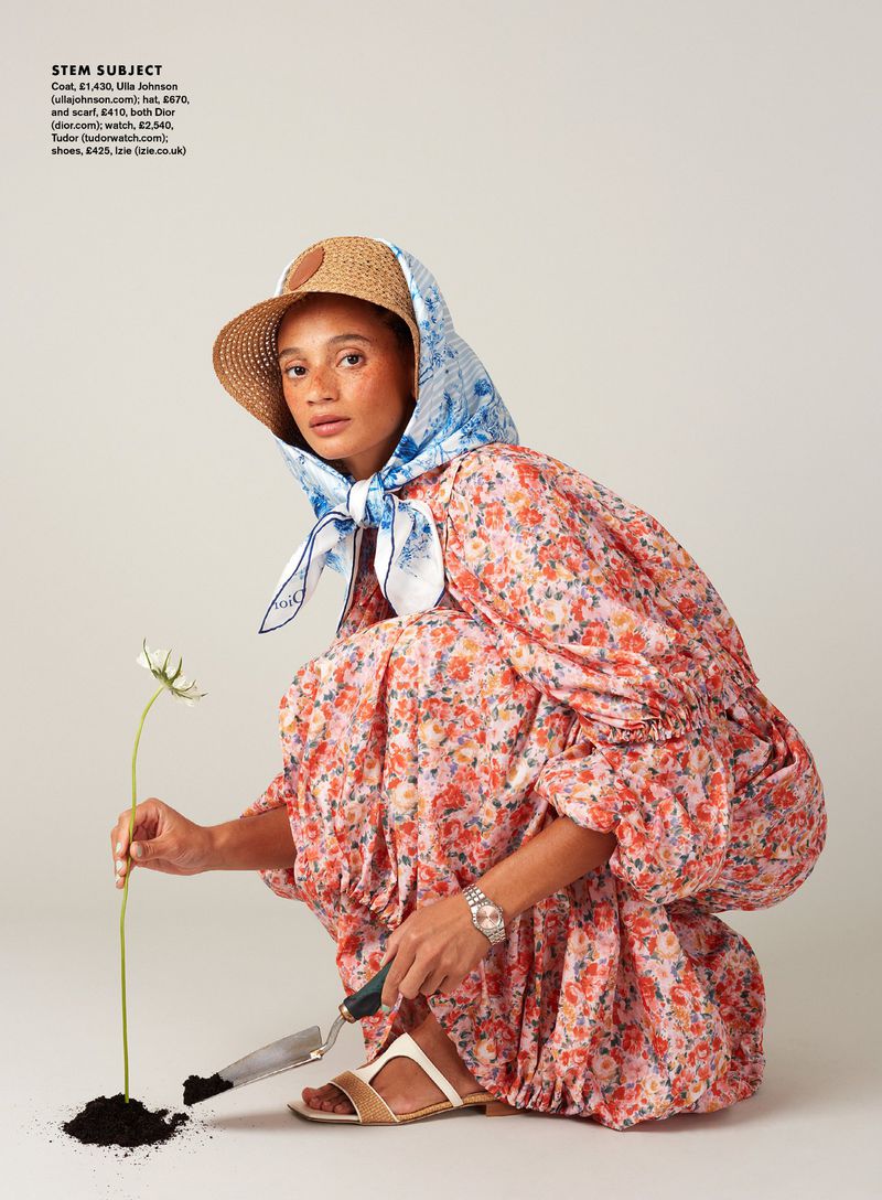 Model Catriona Frean in a pink floral dress and blue silk headscarf crouching with a garden trowel beside a planted flower.