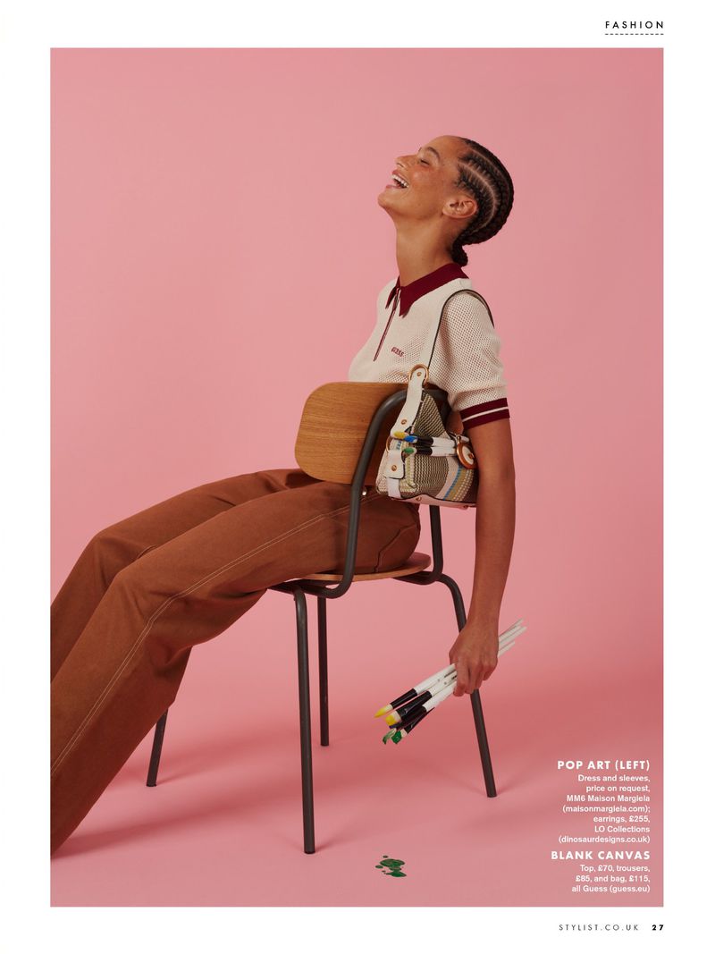 Model Catriona Frean seated on a wooden chair holding paintbrushes against a pink backdrop for Stylist Magazine UK.