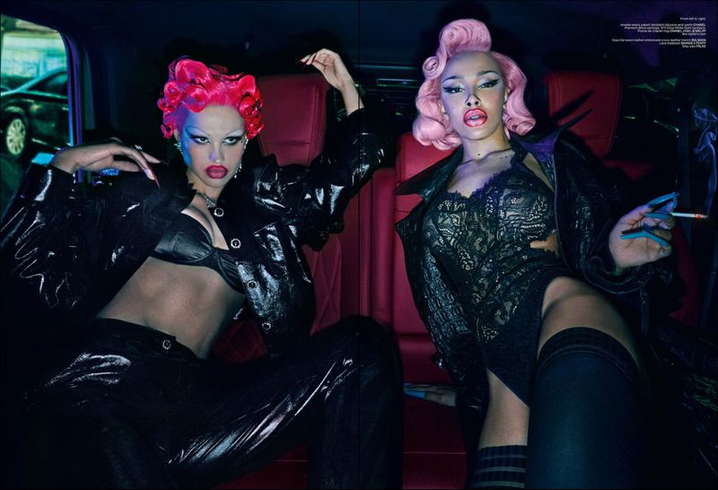 Two women with retro-styled pink hairstyles wearing shiny black outfits pose in the backseat of a car against red leather seats.