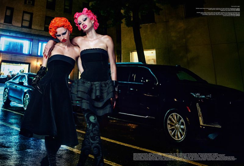 Two women with brightly colored hair pose on a wet city street at night in black dresses, beside a black car under streetlights.