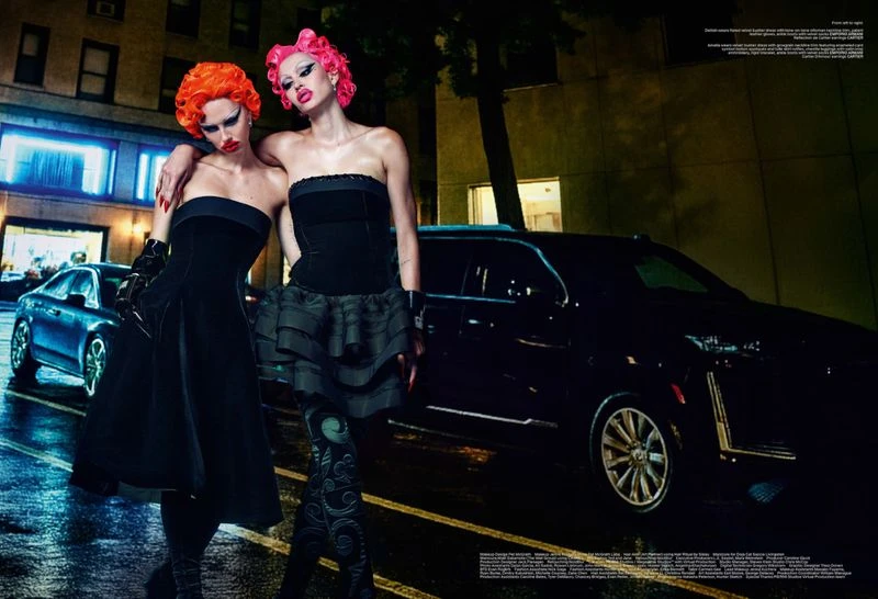 Two women with brightly colored hair pose on a wet city street at night in black dresses, beside a black car under streetlights.