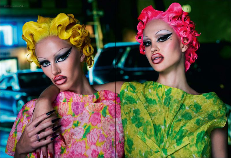 Two women pose in bright floral dresses with vivid pink and yellow curly hair, showcasing bold makeup against an urban backdrop.