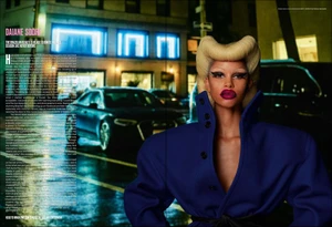 Model Daiane Sodre with sculpted blonde hair wearing an exaggerated royal blue coat on a wet city street at night.