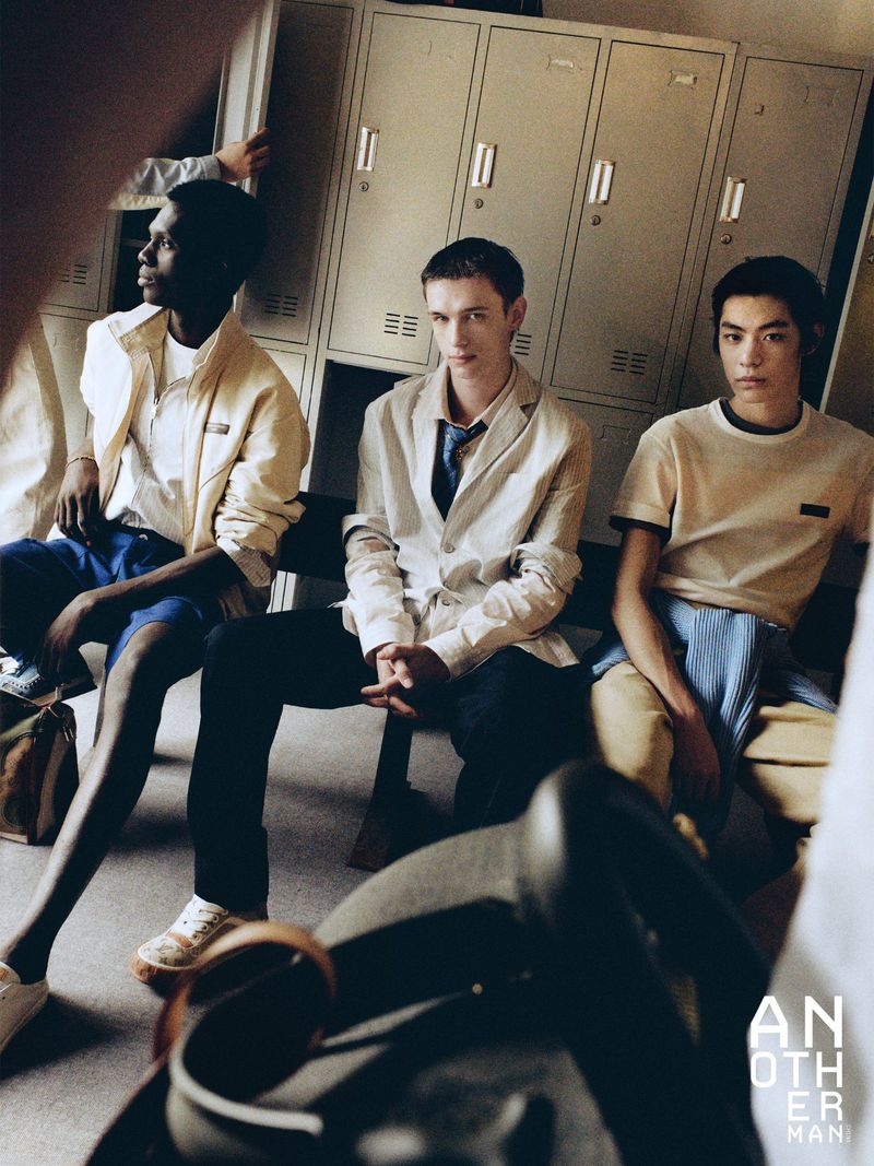 Three male models in casual and tailored menswear seated on a bench in front of vintage metal lockers.