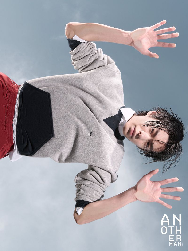 Model Woo Wu posing dynamically in a grey sweatshirt and red shorts against a pale overcast sky for AnOther Man China.