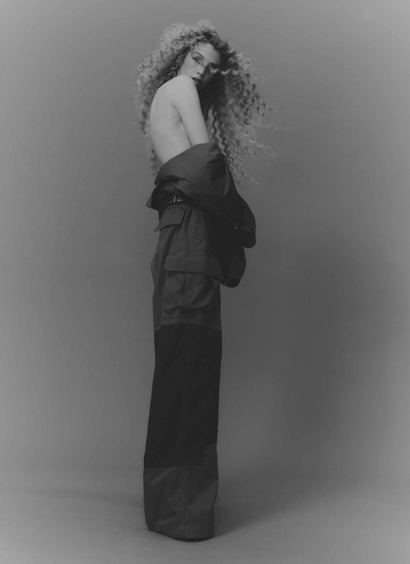 Black and white portrait of a curly-haired woman in wide-leg pants and a jacket draped over her shoulder posing in a studio.