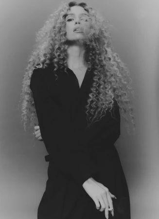 Black and white portrait of a woman with curly hair wearing a long black coat, posing in front of a grey backdrop with a ring on her finger.