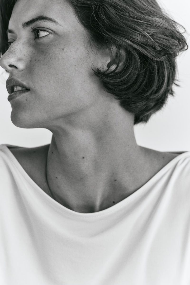 Black and white close-up portrait of Valerie Scherzinger looking profile-left wearing a white boatneck top against a white backdrop.