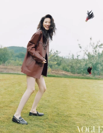 Model Yanan Wan wears a brown stitched leather jacket and loafers while playing shuttlecock on a grass lawn for Vogue China.