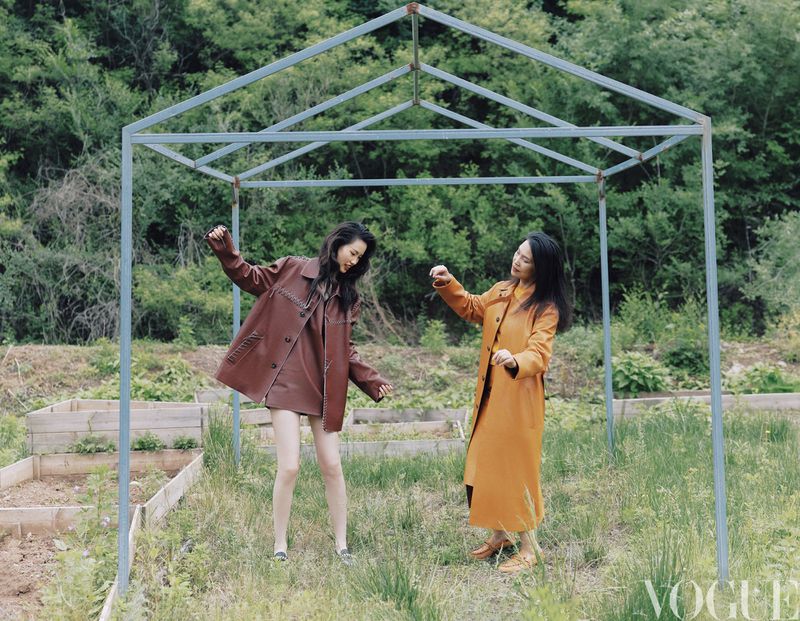 Two East Asian women wearing brown and yellow leather coats pose playfully inside a metal frame structure outdoors.