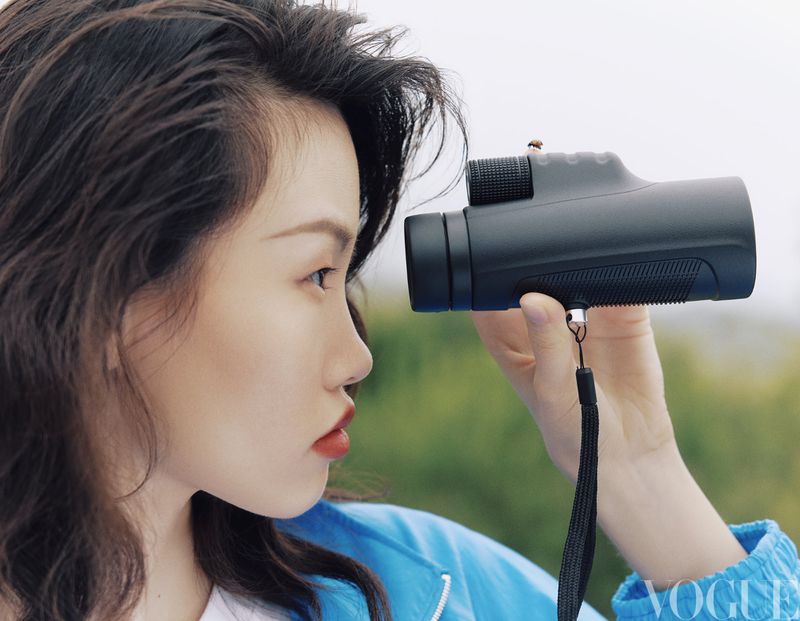 East Asian woman in a blue jacket looking through a black monocular telescope against a soft green outdoor background.