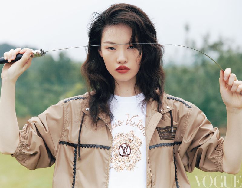 Model Yanan Wan holding a flexible sword horizontally in front of her face while wearing a tan windbreaker jacket outdoors.