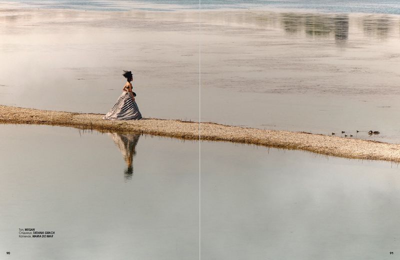 Model Rita Santos wearing a striped sculptural gown while standing on a narrow sandbank in calm water.