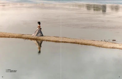 Model Rita Santos wearing a striped sculptural gown while standing on a narrow sandbank in calm water.