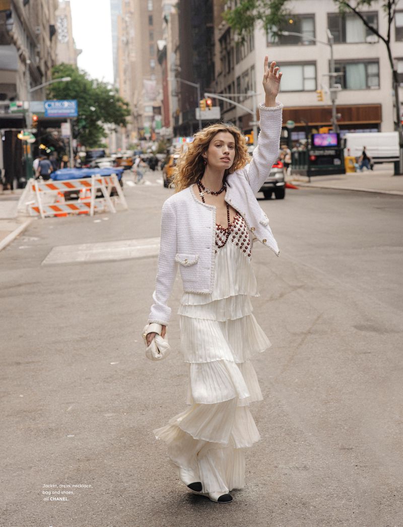 A woman in a white tiered dress stands on a city street with buildings in the background, hailing a taxi with her arm raised.