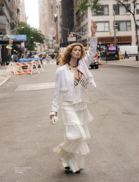 A woman in a white tiered dress stands on a city street with buildings in the background, hailing a taxi with her arm raised.