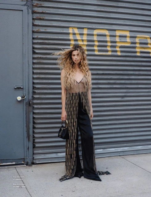 Woman poses against a gray roll-up door wearing a sheer top with fringe, black trousers, heels, and a small black purse.