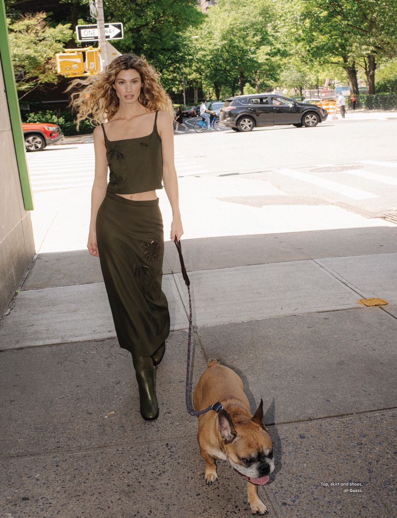 A slender blonde woman walks a tan French bulldog on a city sidewalk, wearing a matching green top, skirt, and boots in natural light.