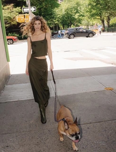 A slender blonde woman walks a tan French bulldog on a city sidewalk, wearing a matching green top, skirt, and boots in natural light.