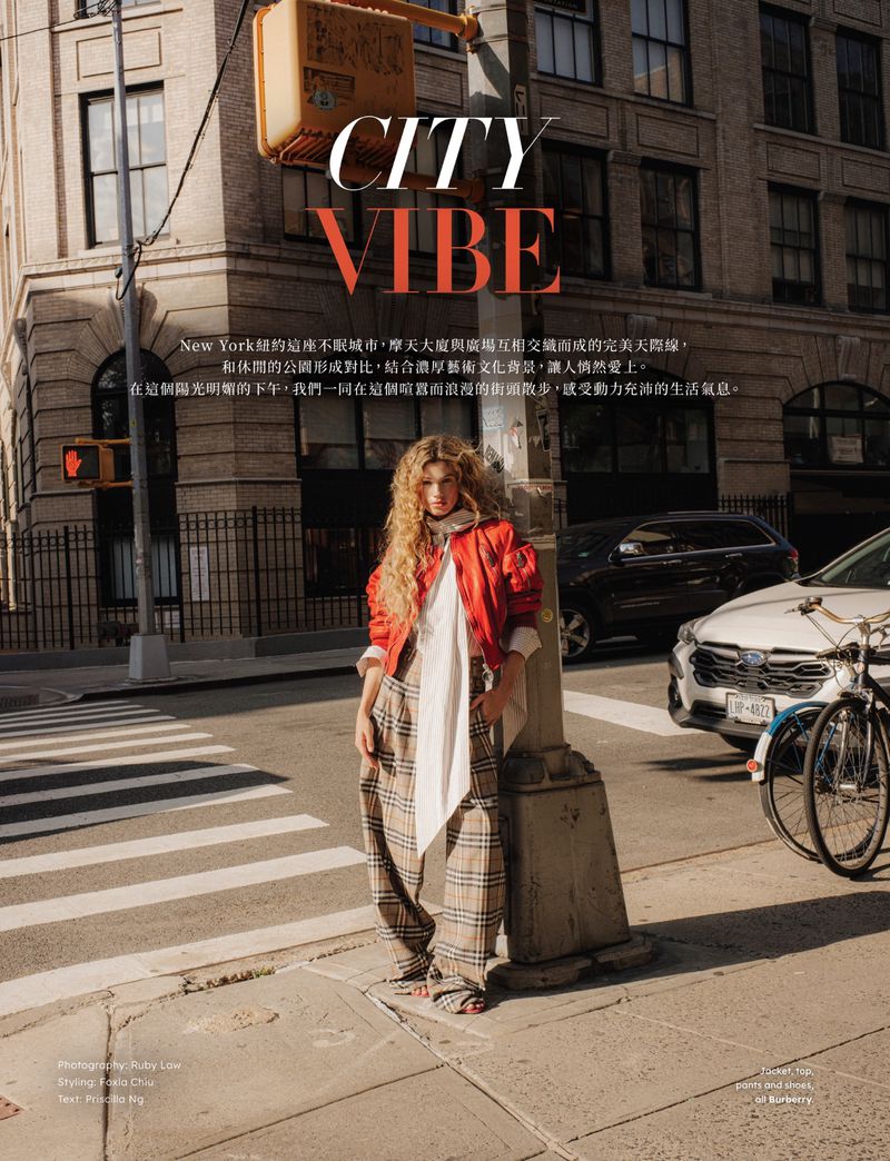 Fashion editorial shot of a model in a red jacket and plaid pants posing on a city street, next to a traffic light pole on a sunny day.