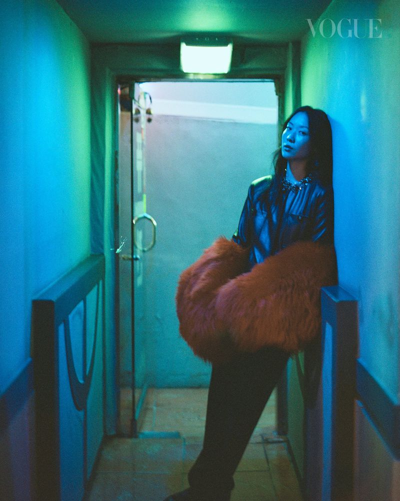 Woman in a dark jacket and oversized orange fur wrap leaning against a wall in a blue and green-lit hallway for Vogue Singapore.