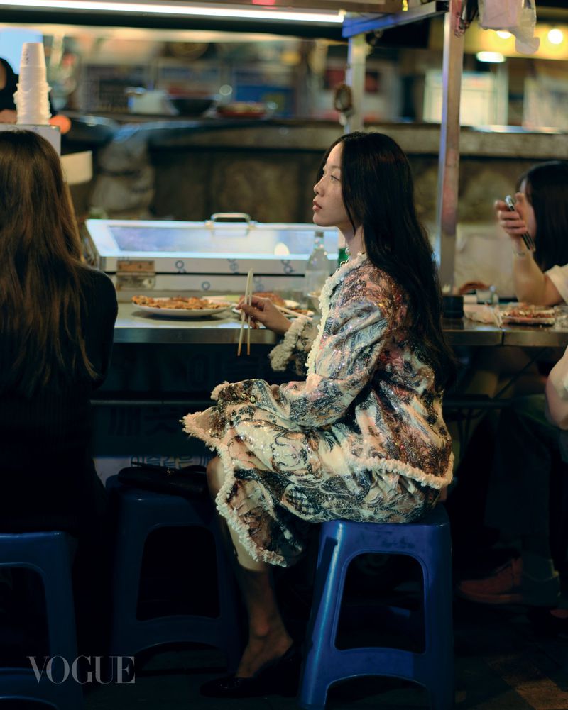East Asian woman in an ornate fringed coat seated on a blue stool at an outdoor food stall holding chopsticks.