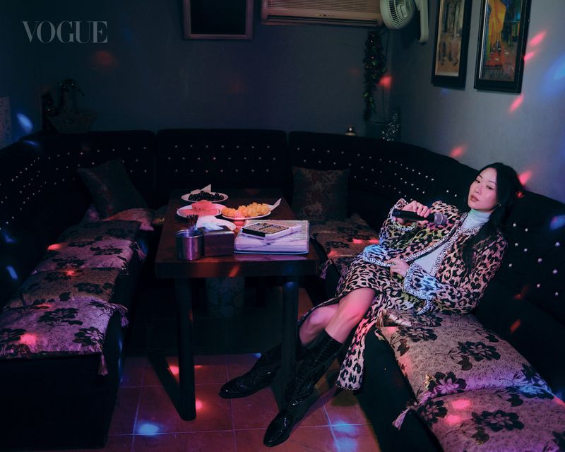Woman in a leopard print coat and black boots reclines on a dark booth holding a microphone under colorful party lights.