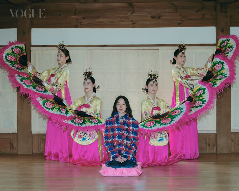 Woman in a blue and red tweed jacket kneeling in front of four Korean fan dancers in pink hanbok holding large feather fans.
