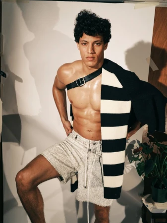 Model Lucas Barski posing shirtless with a leather chest strap, striped sweater draped over shoulder, and textured shorts.