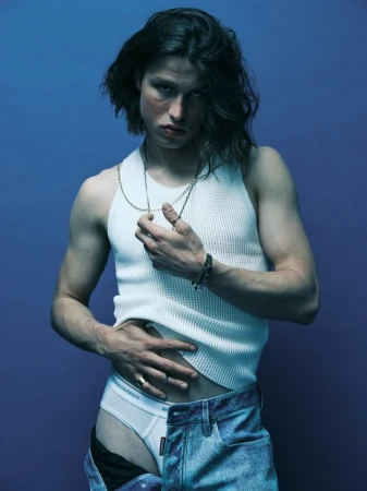 Model Lucas Barski wearing a white ribbed tank top and unbuttoned blue jeans against a solid cool blue studio backdrop.